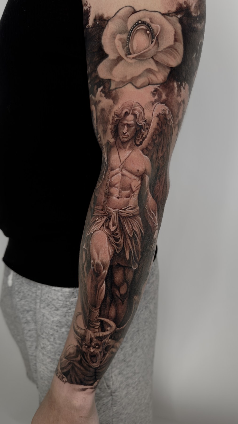 Black and grey arm composition with a rose, winged figure and horned face