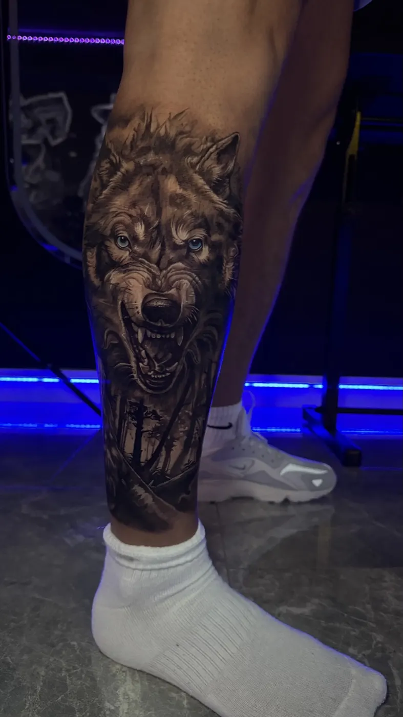 Wolf tattoo on the lower leg with bared teeth, detailed fur and a forest beneath the face — by Laurent Alba
